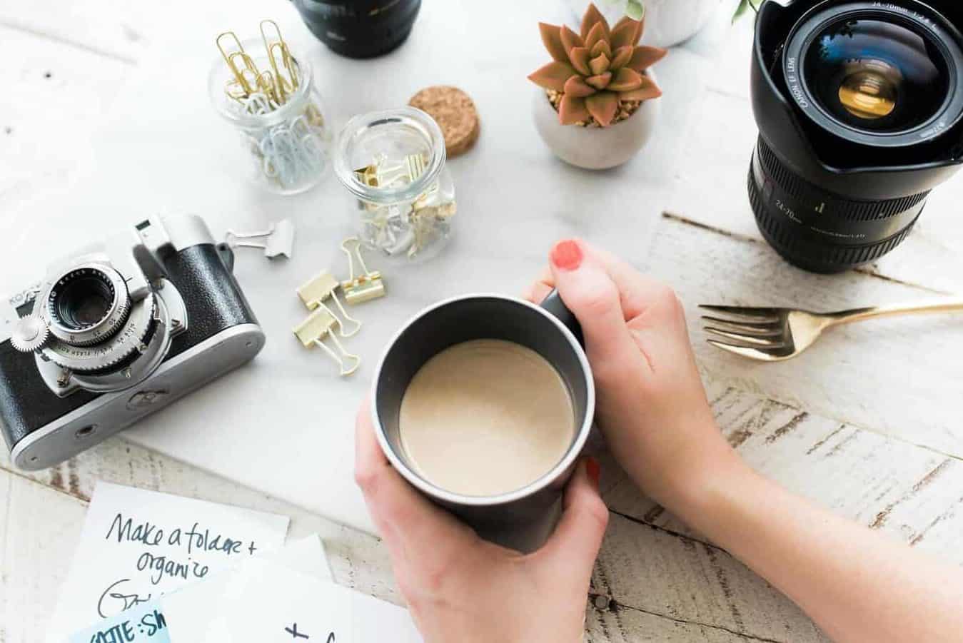 Tasse Kaffee mit Hafermilch, von Händen gehalten, auf Tisch, drum herum Kamera, Objektiv und Gläser mit Papierclips