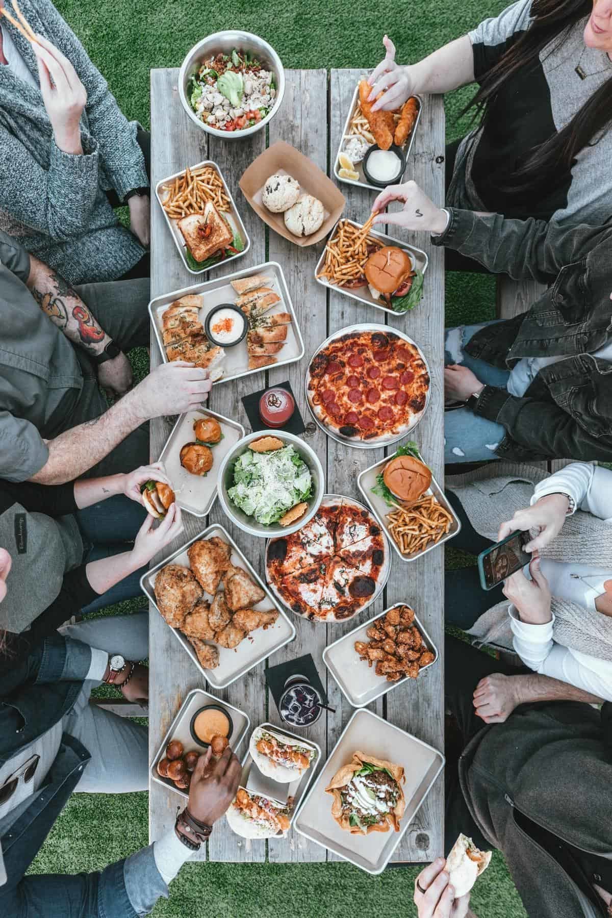 Vegane Speisen in Schüsseln auf Tisch, drum herum Menschen, die zugreifen und fotografieren